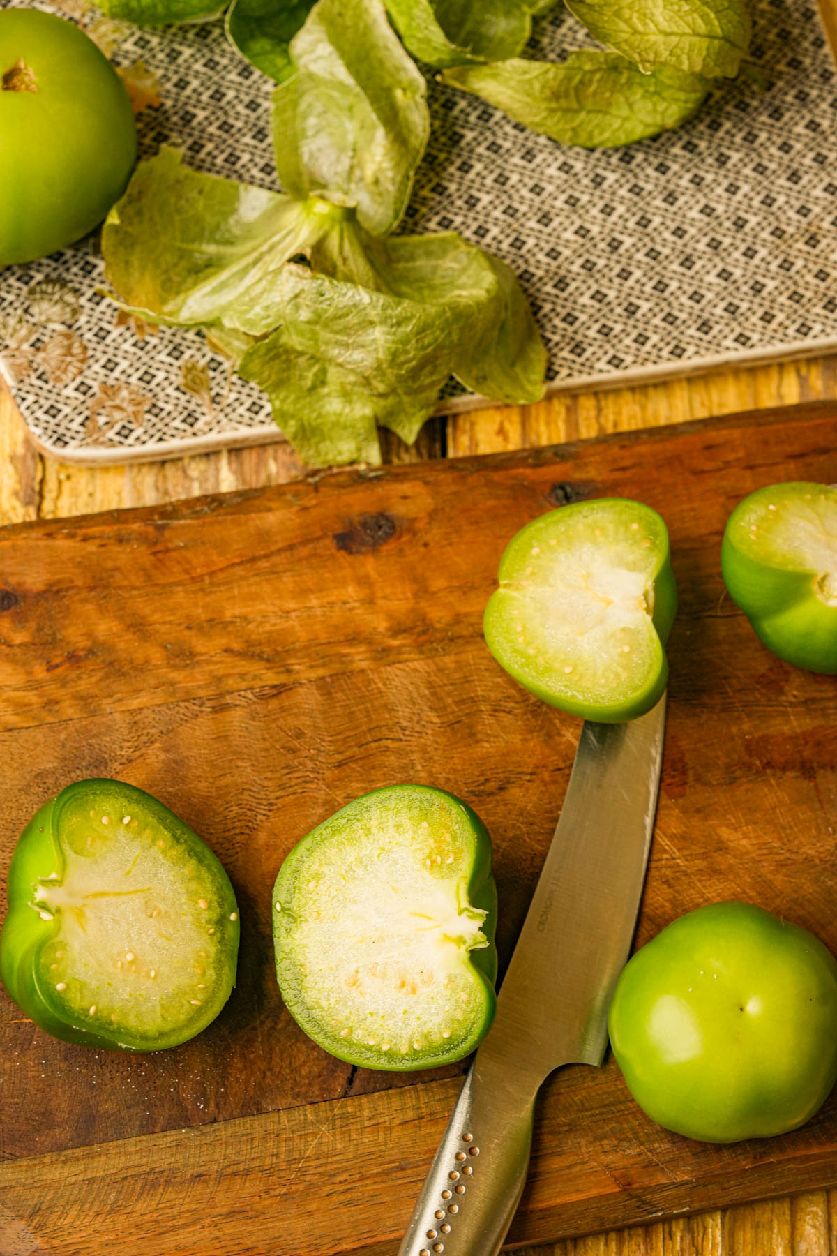 Sliced green tomatillos and a knife on a wooden cutting board, with fresh green tomatillo husks on a patterned cloth in the background.