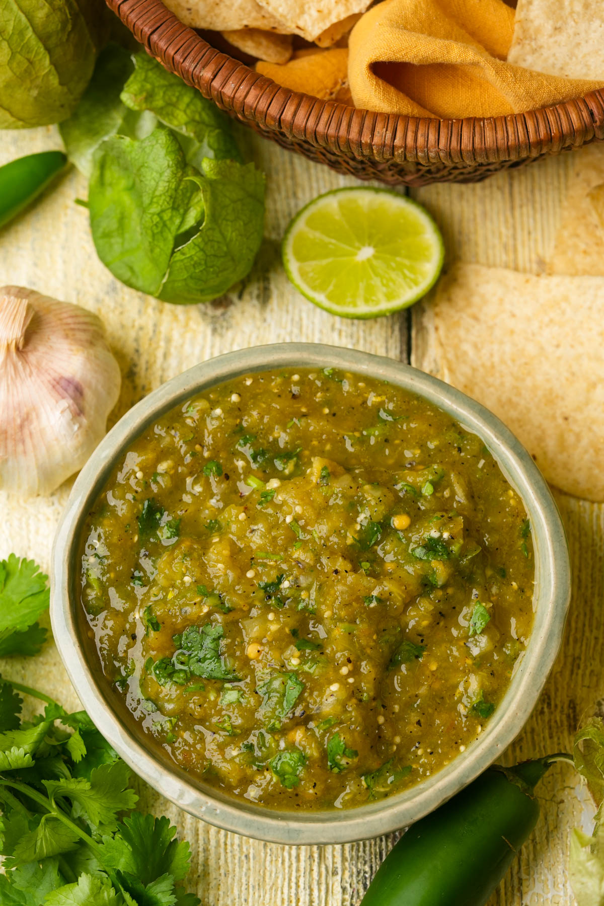 A bowl of hatch chile salsa with cilantro sits on a wooden surface surrounded by a halved lime, a garlic bulb, tomatillos, a jalapeño, cilantro leaves, and a basket of tortilla chips.