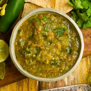 A bowl of hatch chile salsa with cilantro, surrounded by tortilla chips, a whole jalapeño, lime, and fresh cilantro on a wooden surface.