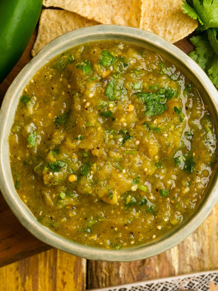 A bowl of hatch chile salsa with cilantro, surrounded by tortilla chips, a whole jalapeño, lime, and fresh cilantro on a wooden surface.