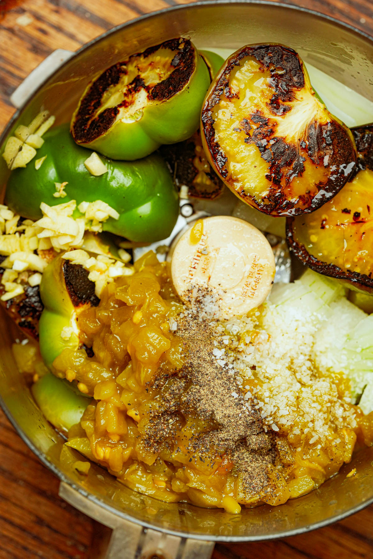 A food processor bowl containing charred green tomatillos, chopped garlic, chopped onions, spices, and canned hatch chilies ready to be blended.