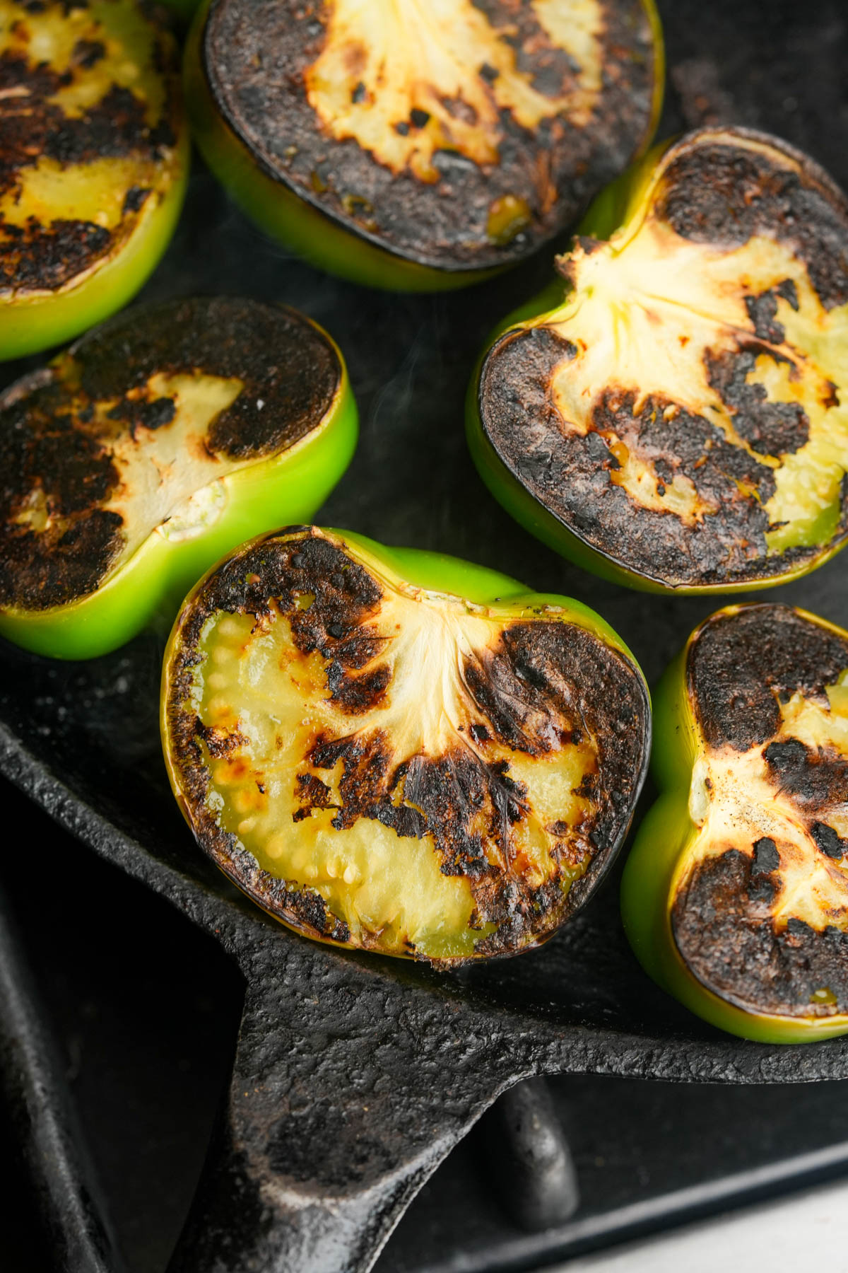 Halved green tomatillos with charred, blackened tops grilling on a cast iron skillet. The edges are browned, and the skillet is on a stovetop.
