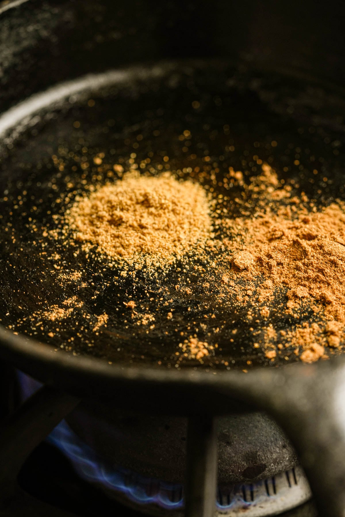 pPles of ground cumin and coriander are toasting in a black cast iron skillet on a stove, with a faint blue gas flame visible underneath.
