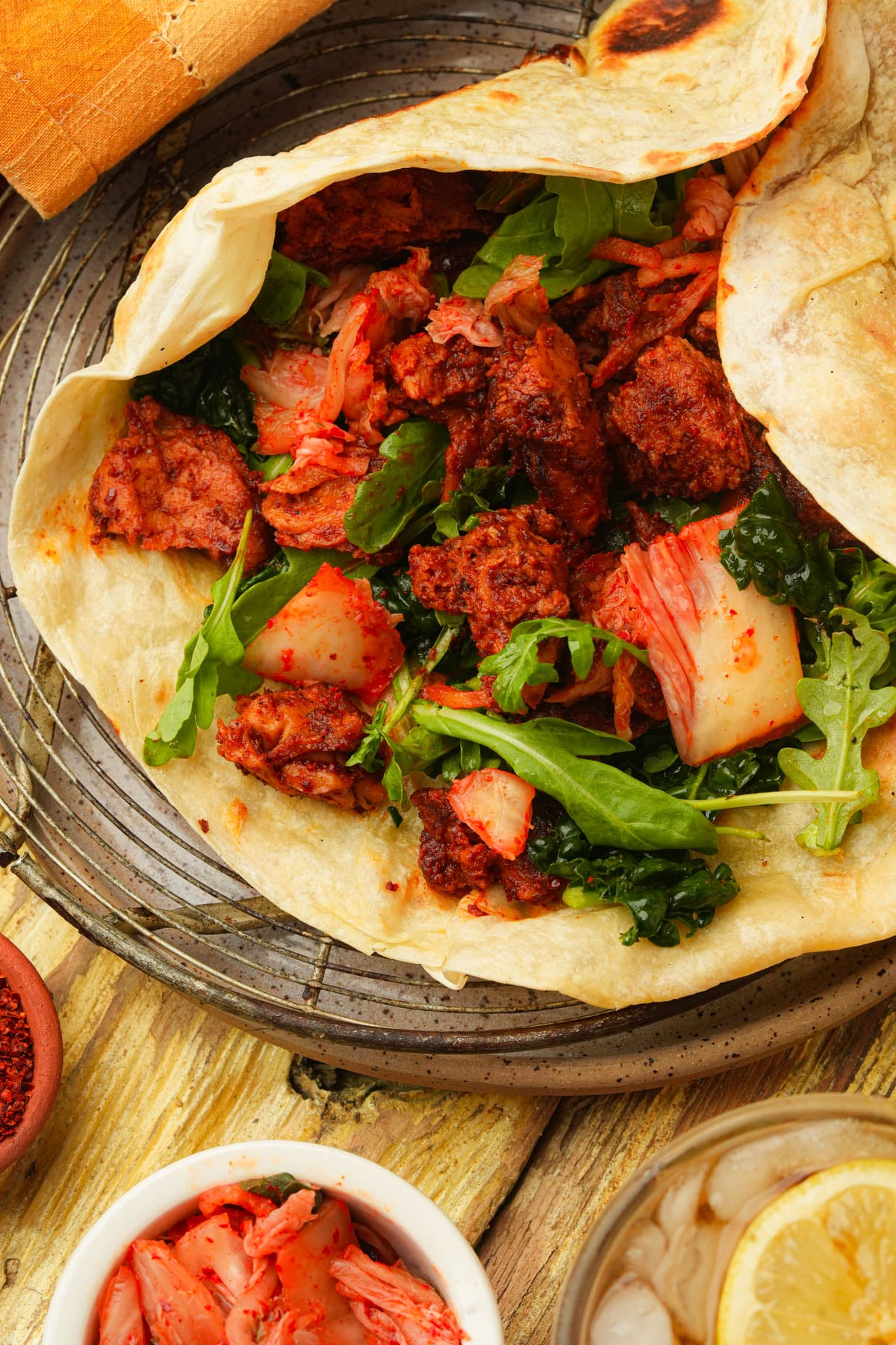 A close-up of Korean BBQ seitan filled with seasoned seitan, kimchi, arugula, and leafy greens on a wire rack, with a small bowl of kimchi and a glass of ice water with lemon nearby.