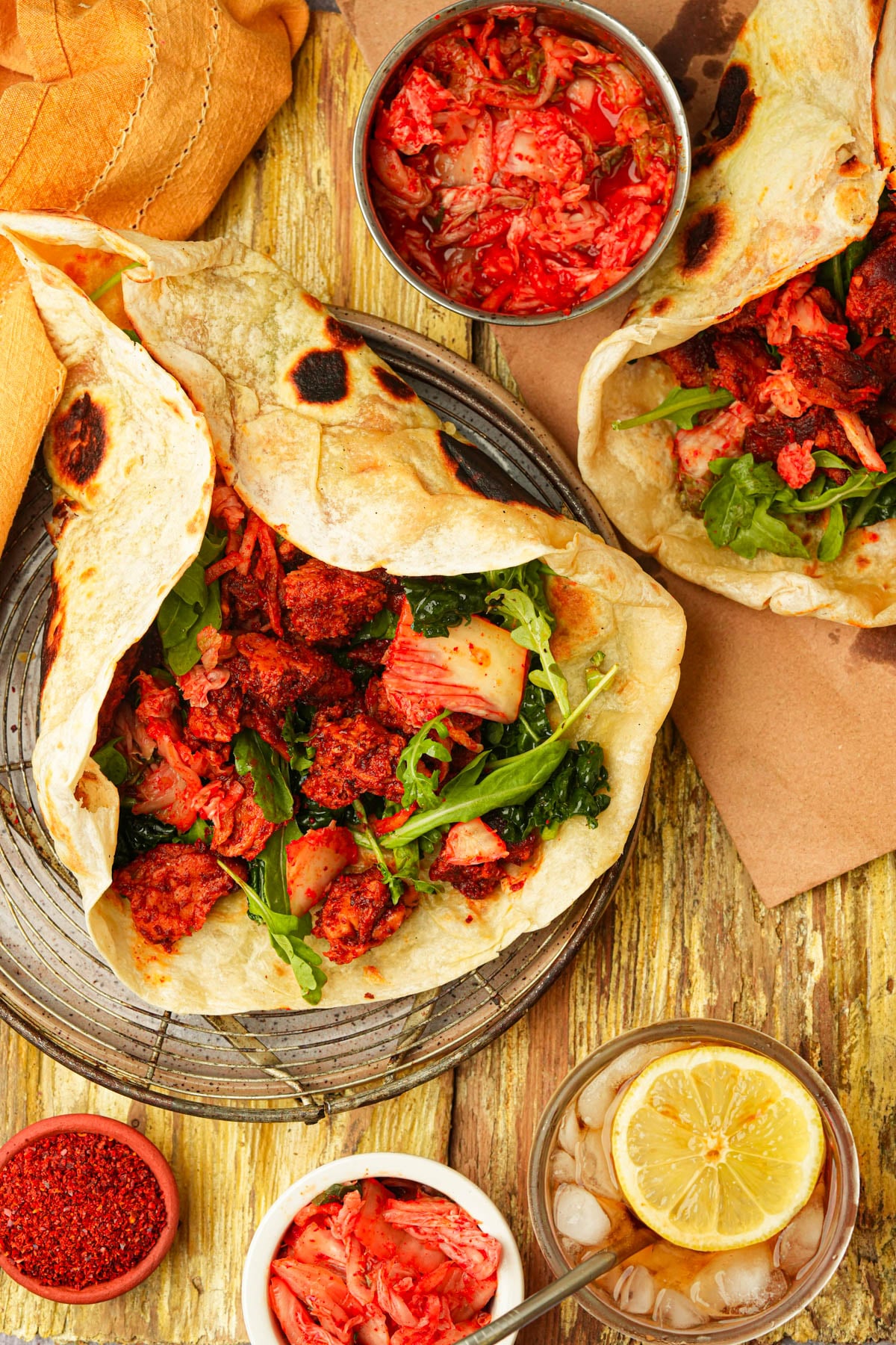 Two orders of Korean BBQ seitan filled with greens and spicy red seitan are served on a wooden table, accompanied by bowls of kimchi, ice water with lemon, and a small dish of red spice.