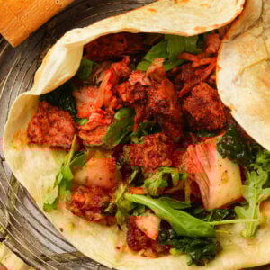 A grilld tortilla filled with spicy Korean BBQ seitan, kimchi, fresh arugula, and leafy greens, placed on a wire rack with a piece of bread visible in the corner.