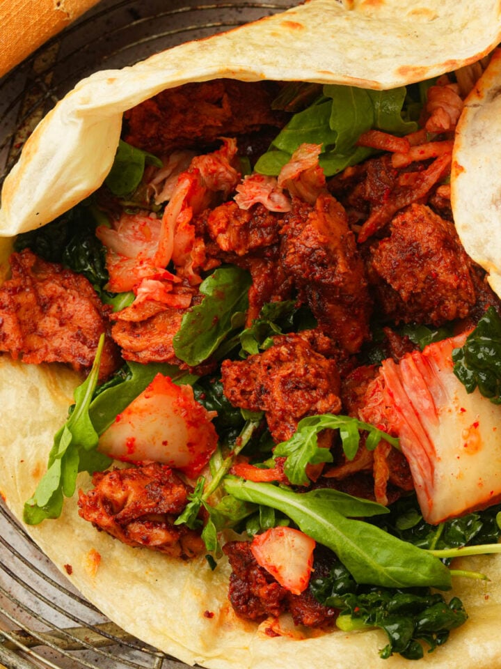 A grilld tortilla filled with spicy Korean BBQ seitan, kimchi, fresh arugula, and leafy greens, placed on a wire rack with a piece of bread visible in the corner.