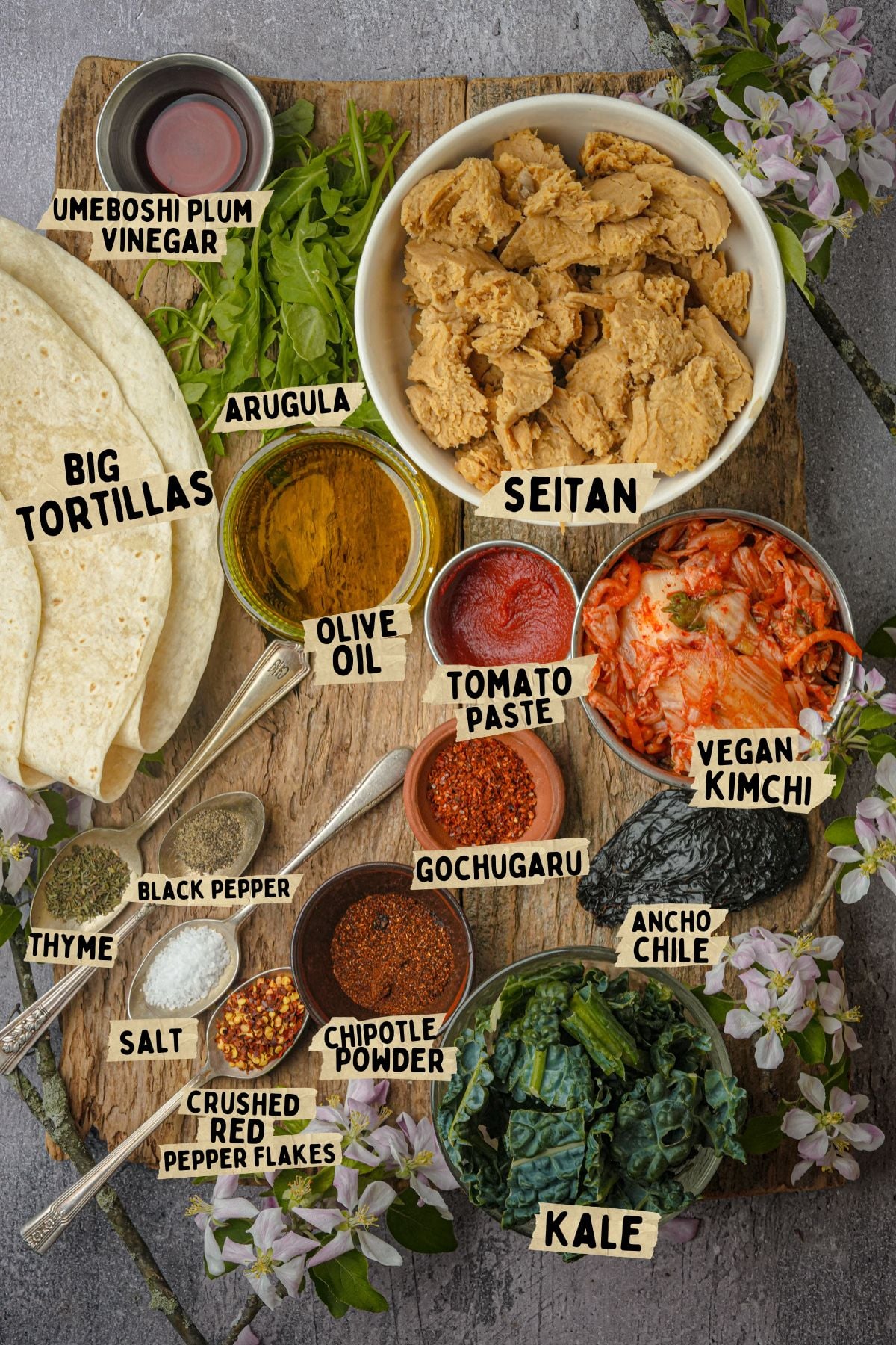 A wooden board holds labeled ingredients for a Korean BBQ seitan recipe: big tortillas, umeboshi plum vinegar, arugula, seitan, olive oil, tomato paste, vegan kimchi, gochugaru, ancho chile, kale, chipotle powder, thyme, black pepper, salt, and red pepper flakes.