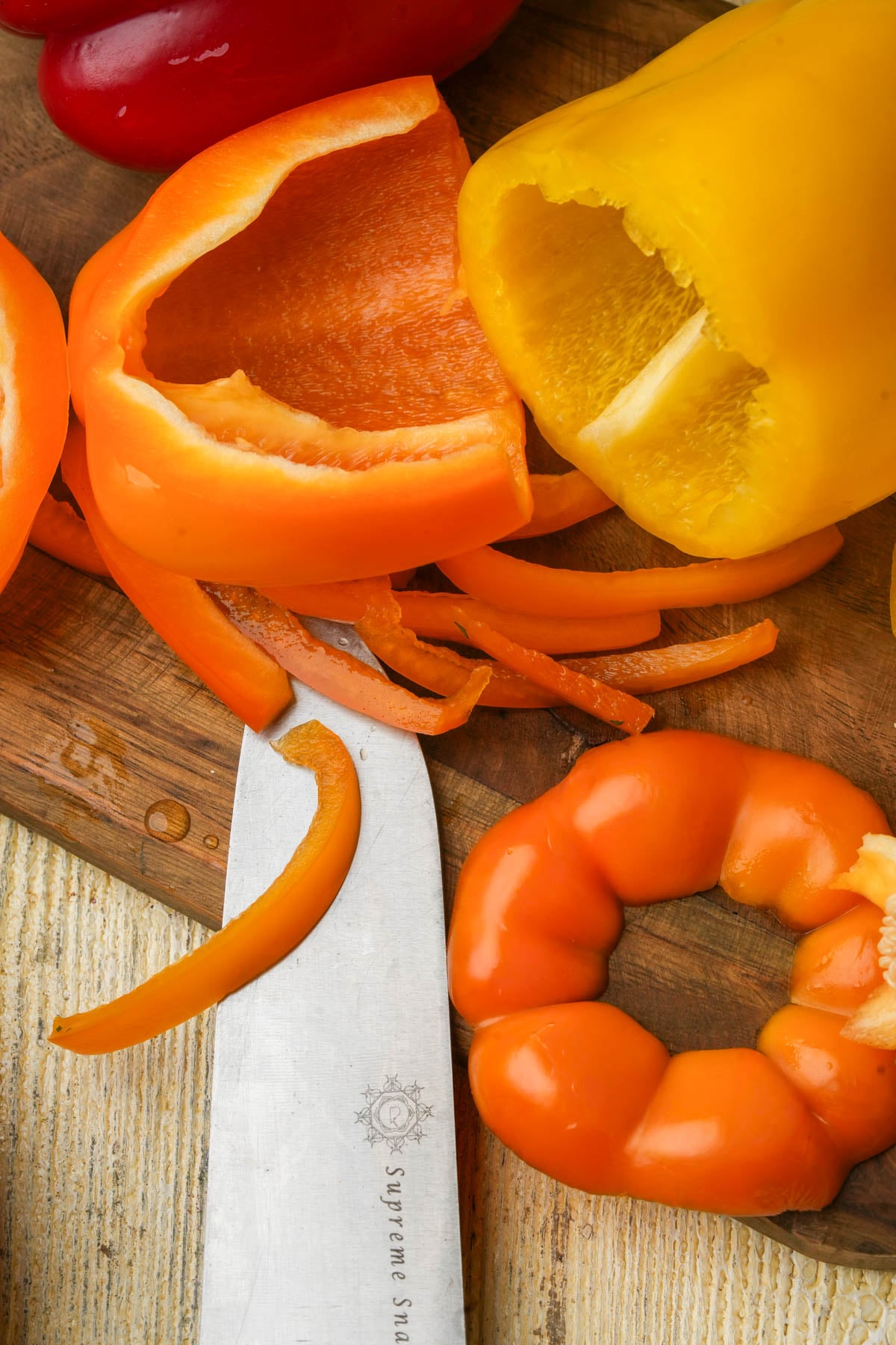 Sliced orange and yellow bell peppers on a wooden cutting board, with a knife nearby. Seeds and pepper tops are also visible, showing food preparation in progress.