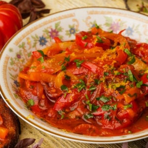 A bowl of stewed peperonata garnished with chopped herbs, sits on a floral-patterned plate.