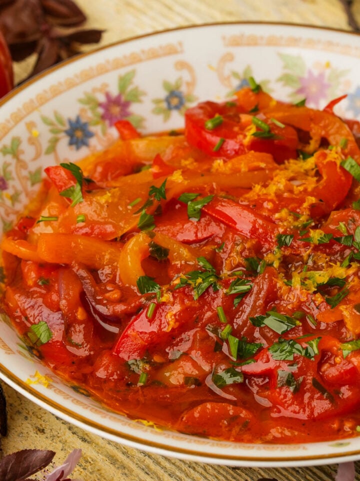 A bowl of stewed peperonata garnished with chopped herbs, sits on a floral-patterned plate.