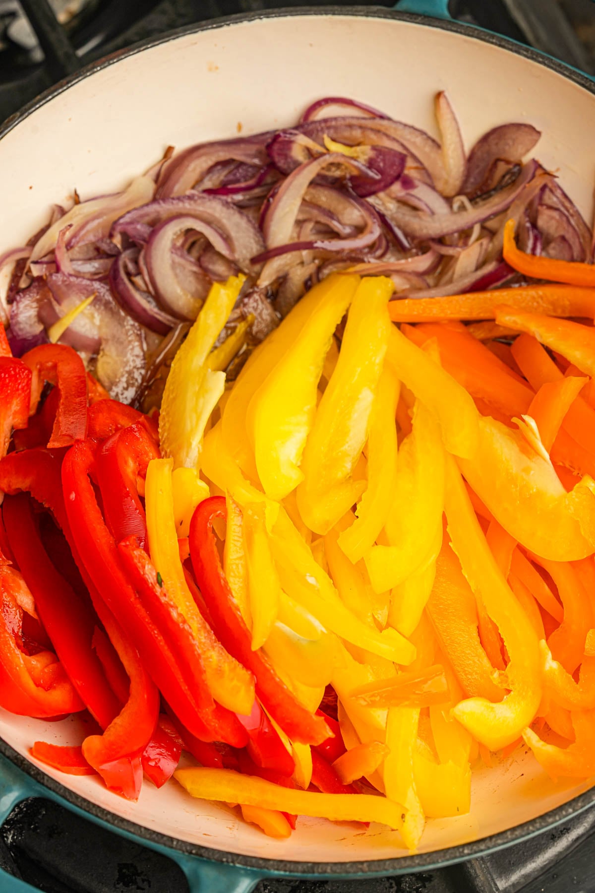 Sliced red onions, red bell peppers, yellow bell peppers, and orange bell peppers cooking together in a white skillet on a stovetop.