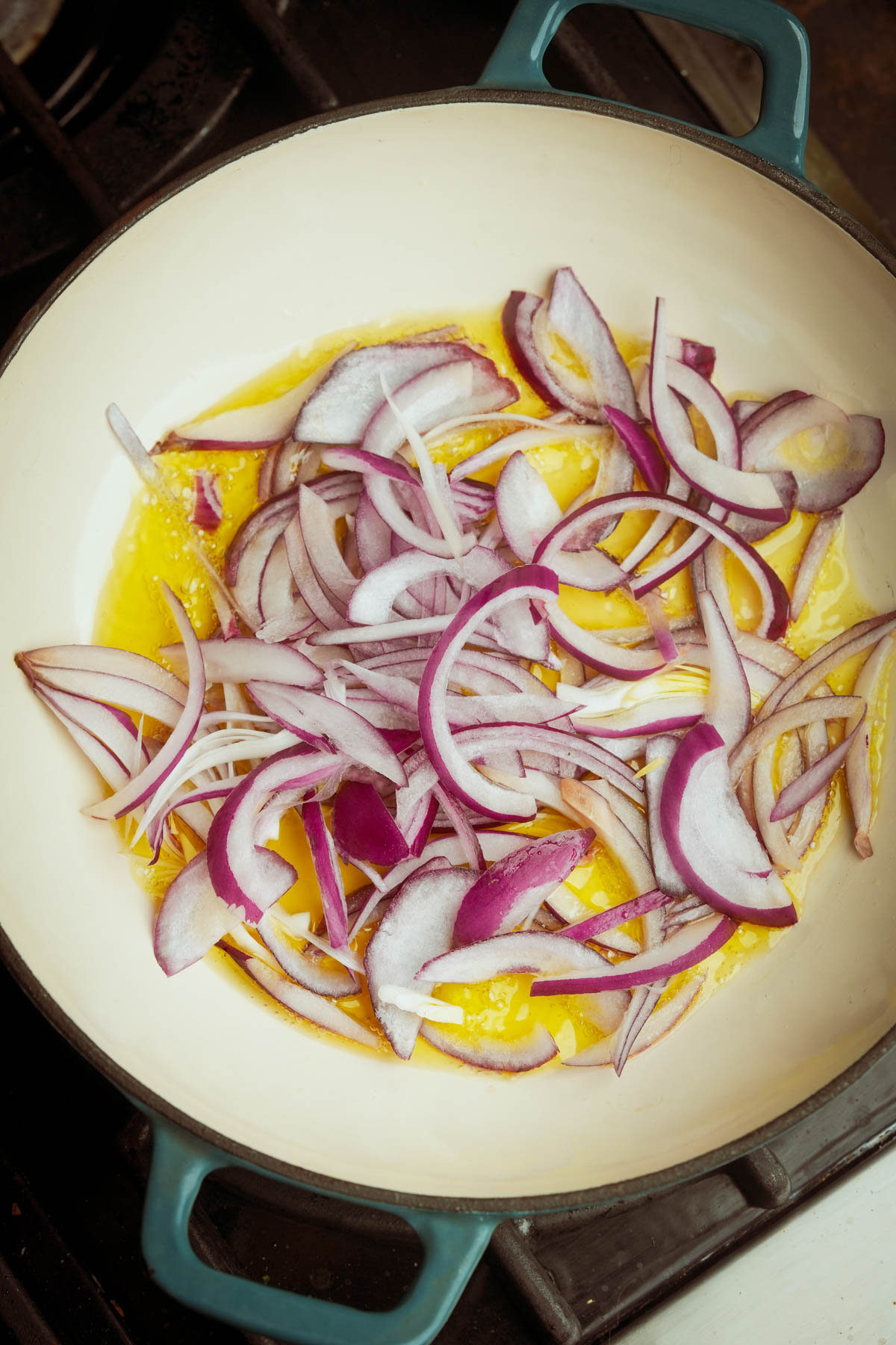 Sliced red onions cooking in a white pot with olive oil on a stovetop, starting to soften and sauté.