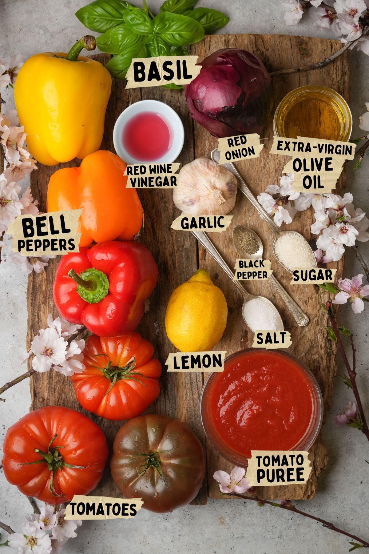 A wooden board topped with ingredients for a peperonata recipe including fresh basil, yellow and red bell peppers, red onion, olive oil, red wine vinegar, garlic, black pepper, sugar, lemon, salt, various tomatoes, and a bowl of tomato puree.