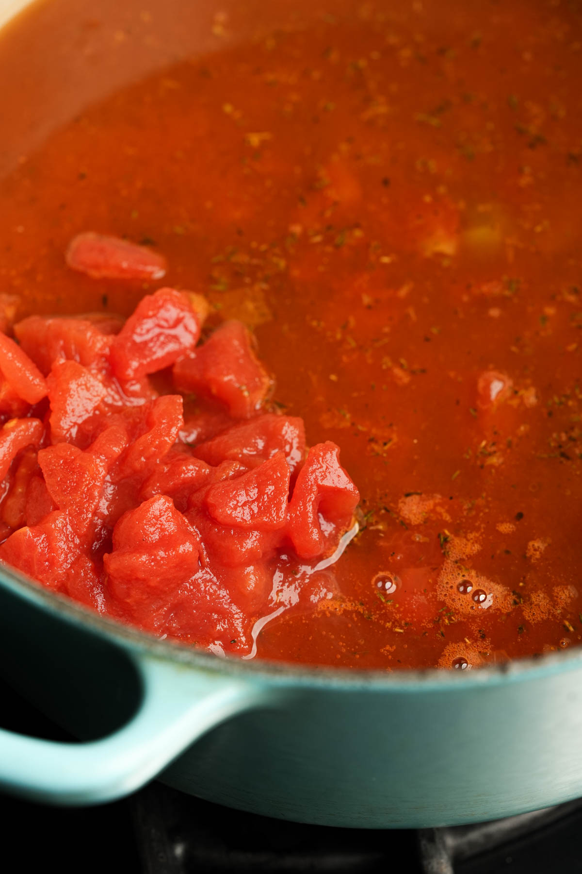 Tomatoes being added to a pot of simmering vegan minestrone in a light blue enameled pot on a stovetop.