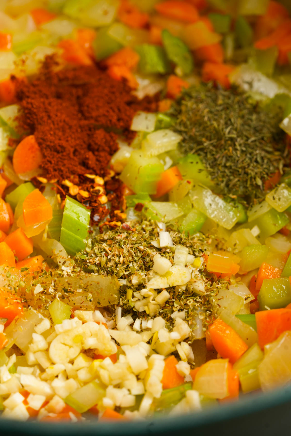 Close-up of chopped carrots, celery, onions, and garlic in a pan, topped with dried herbs, red chili flakes, and paprika, ready to be cooked.