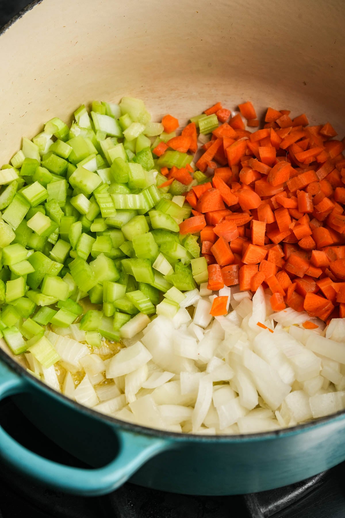 Chopped celery, carrots, and onions are evenly arranged in sections in a large pot on a stovetop, ready to be sautéed.