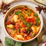 A bowl of vegan minestrone soup with pasta shells, kidney beans, green beans, tomatoes, carrots, celery, and parsley, served on a plate with a spoon and garnished with fresh herbs.