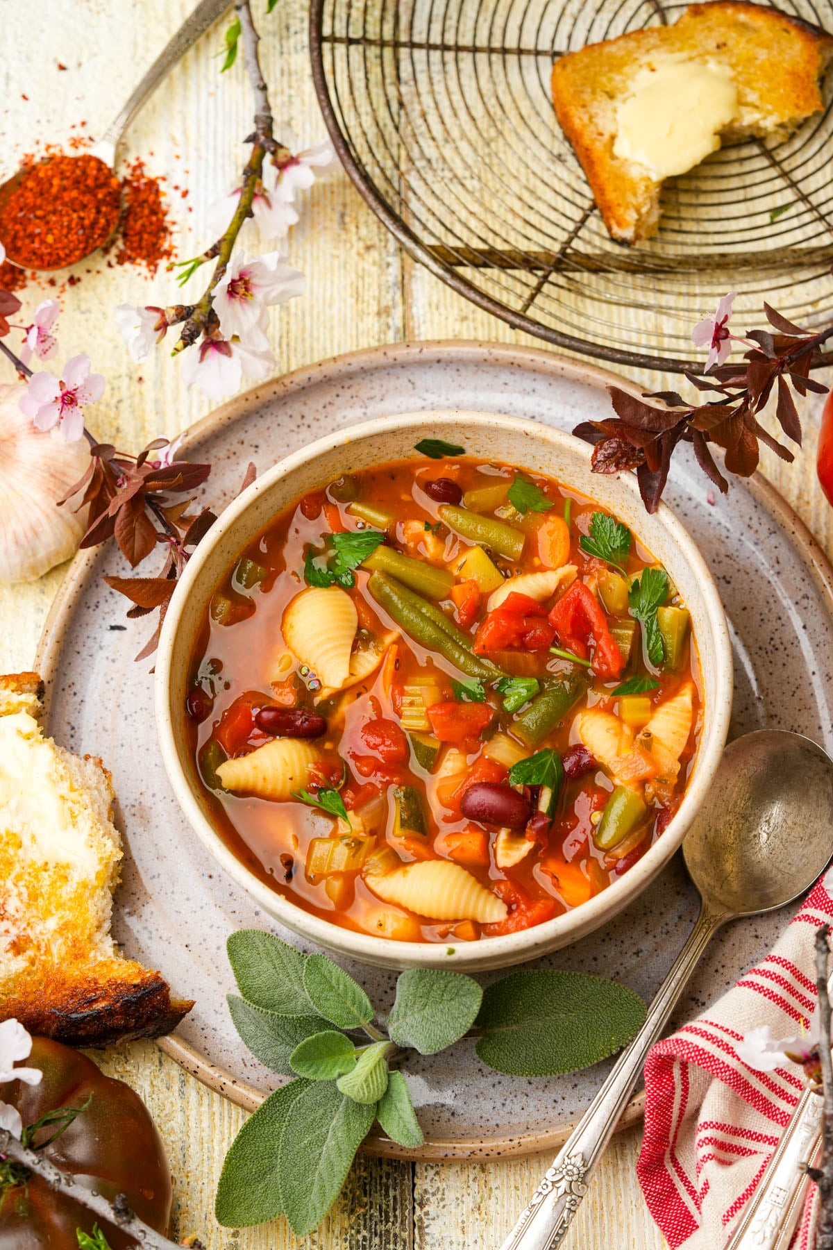 A bowl of vegan minestrone soup with pasta shells, tomatoes, green beans, and kidney beans sits on a plate beside a spoon, herbs, and buttered toast on a rustic wooden table.