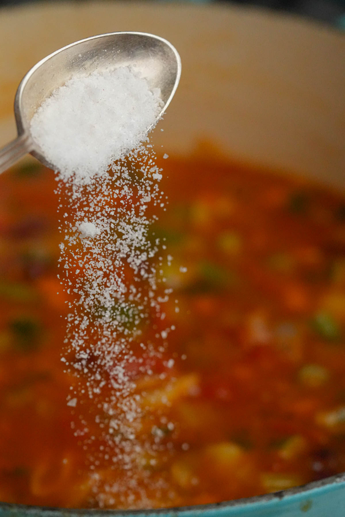 A spoon pours salt into a pot of colorful vegan minestrone soup with visible vegetables, adding seasoning during cooking.