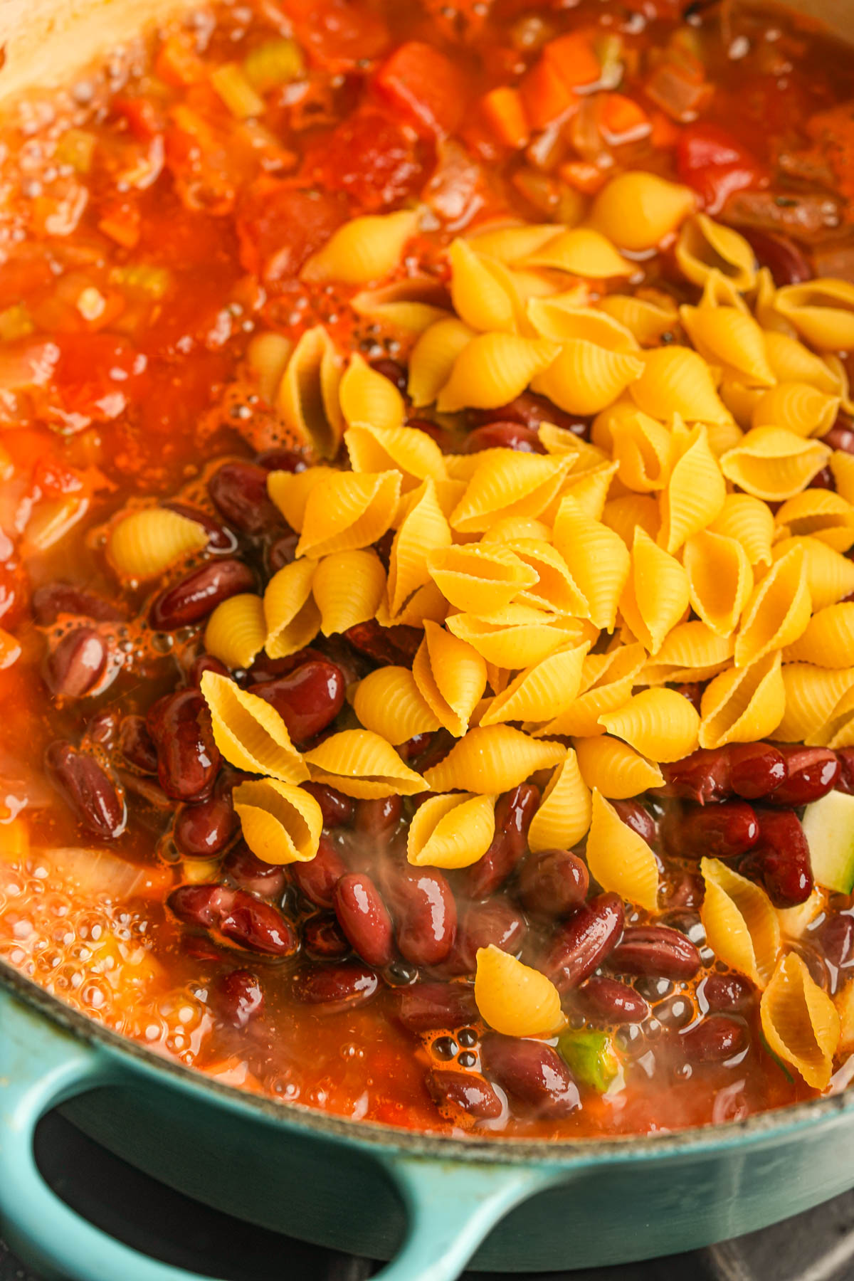 A pot of vegan minestrone is cooking, filled with red kidney beans, chopped vegetables, tomato broth, and uncooked small shell pasta on top, ready to be mixed in.