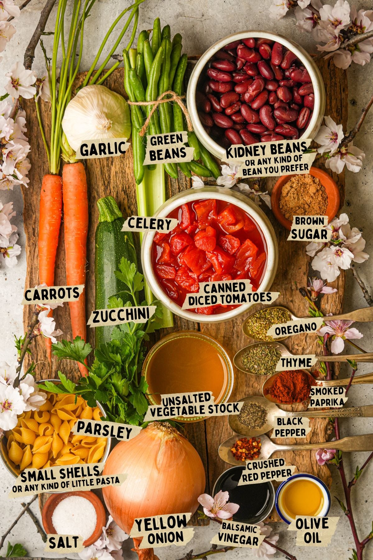 A wooden board displays labeled ingredients for a vegan minestrone soup recipe including carrots, garlic, green beans, kidney beans, celery, zucchini, parsley, small shells pasta, yellow onion, canned diced tomatoes, vegetable stock, spices, oil, and vinegar.