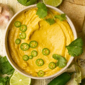 A bowl of creamy vegan queso is garnished with sliced green jalapeños and fresh cilantro. Surrounding the bowl are lime halves, cilantro leaves, a whole jalapeño, and a bulb of garlic.