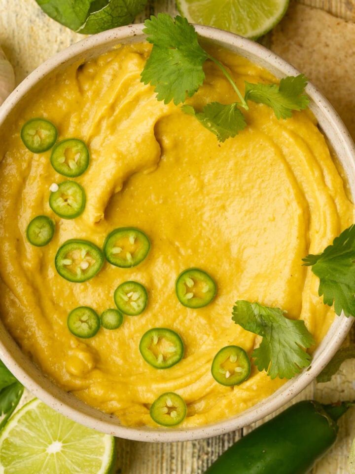 A bowl of creamy vegan queso is garnished with sliced green jalapeños and fresh cilantro. Surrounding the bowl are lime halves, cilantro leaves, a whole jalapeño, and a bulb of garlic.