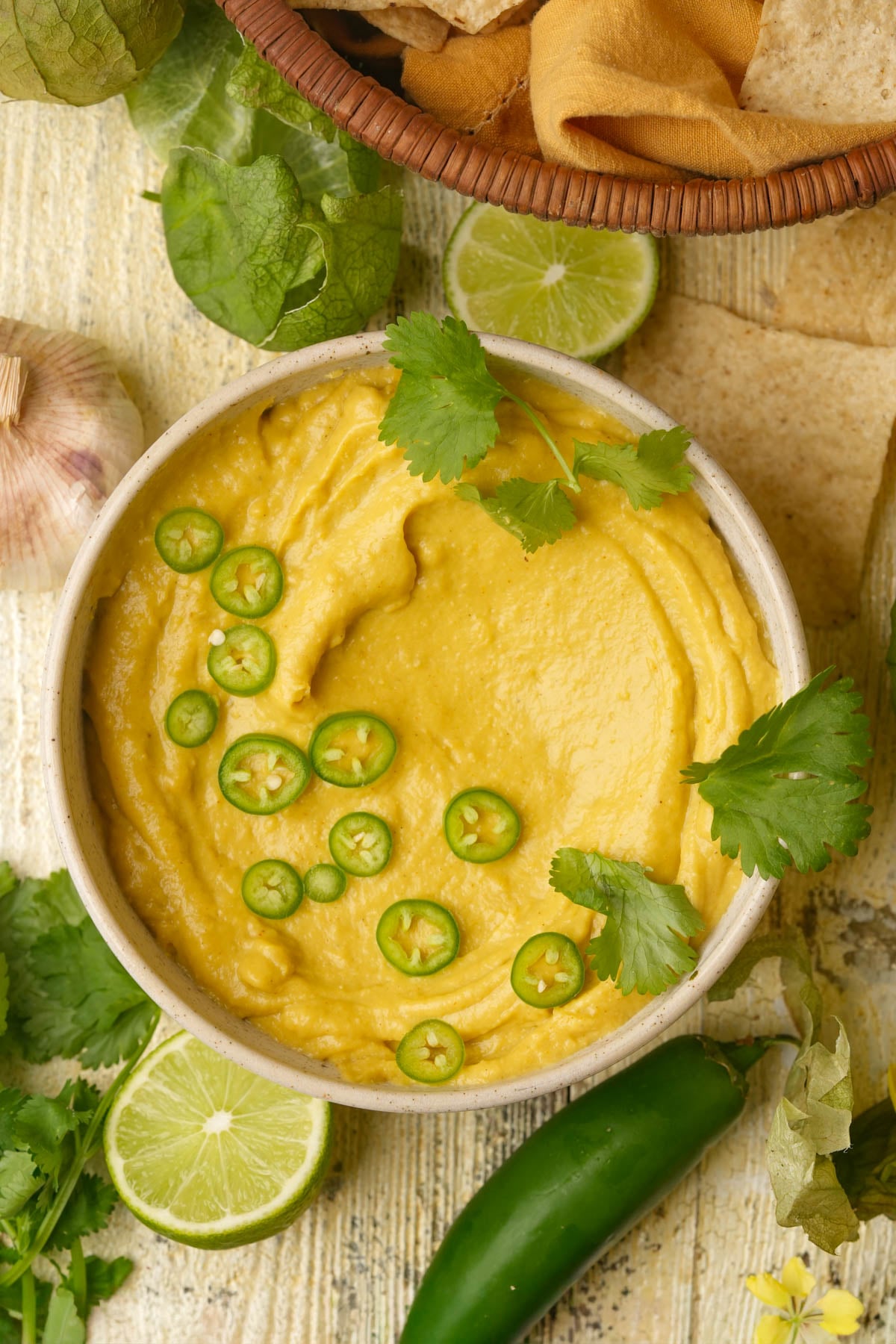 A bowl of vegan queso topped with cilantro leaves and sliced green chili peppers, surrounded by lime halves, garlic, fresh greens, and tortilla chips on a rustic surface.