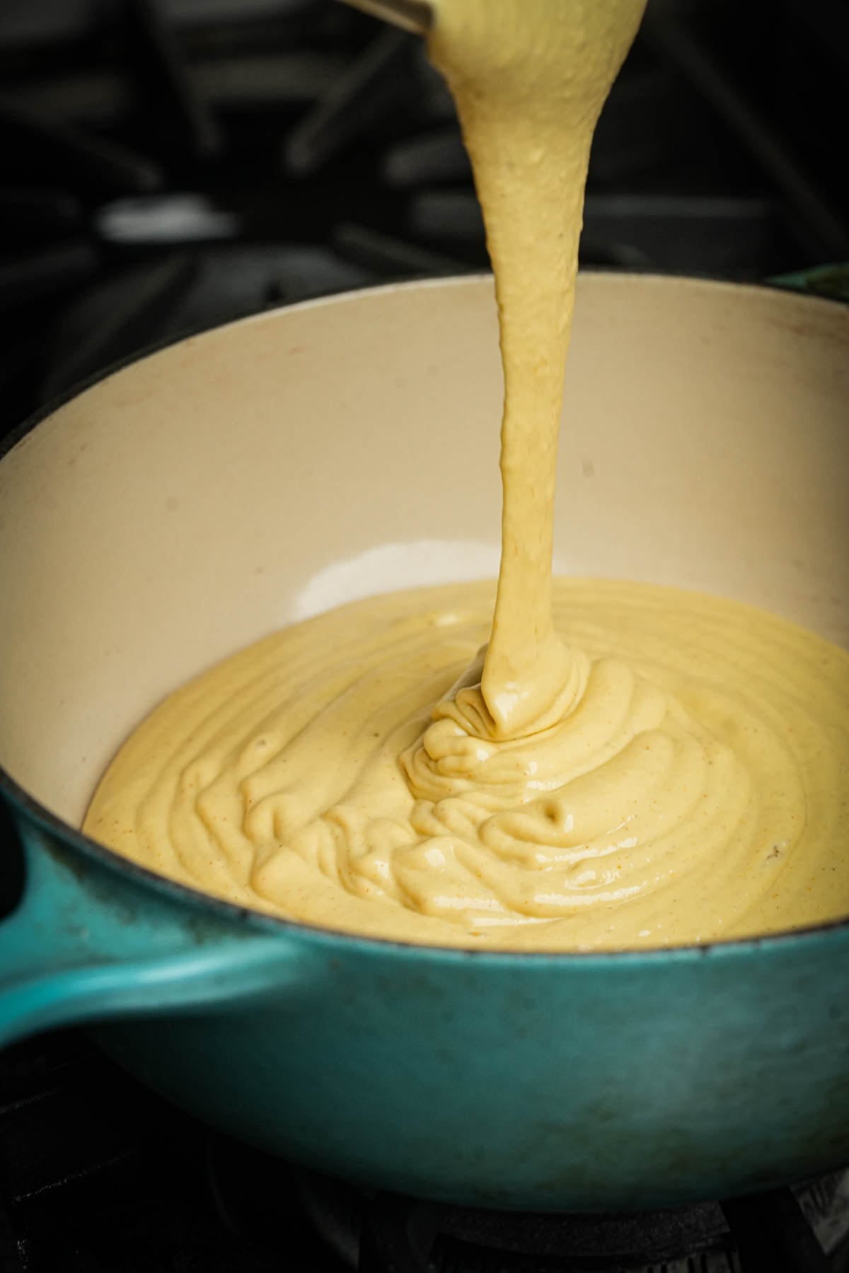 Pureed vegan queso is being poured into a turquoise Dutch oven on a stovetop, creating swirls as it spreads out inside the pot.