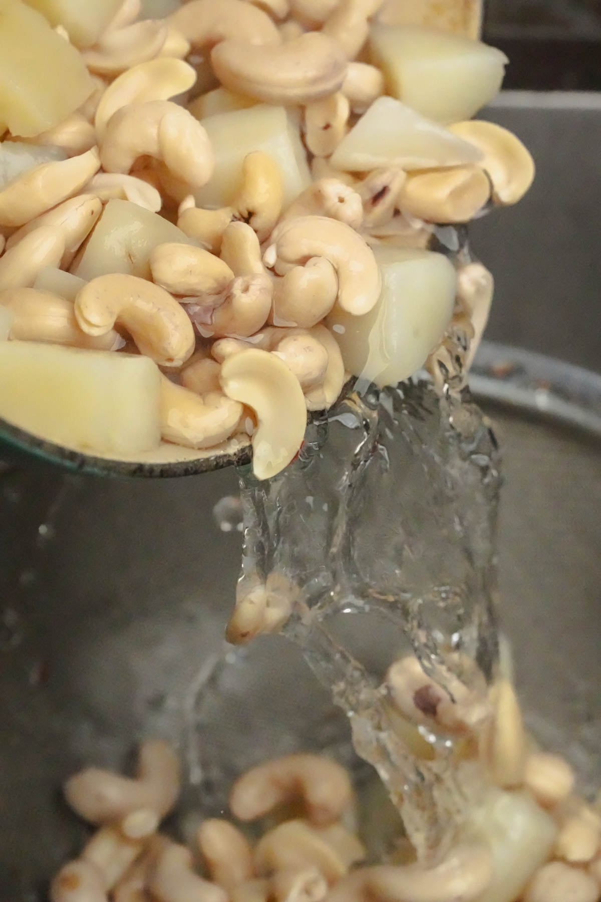 Cashew nuts and potato pieces being poured with water into a strainer, with water splashing over the cashews and potatoes.