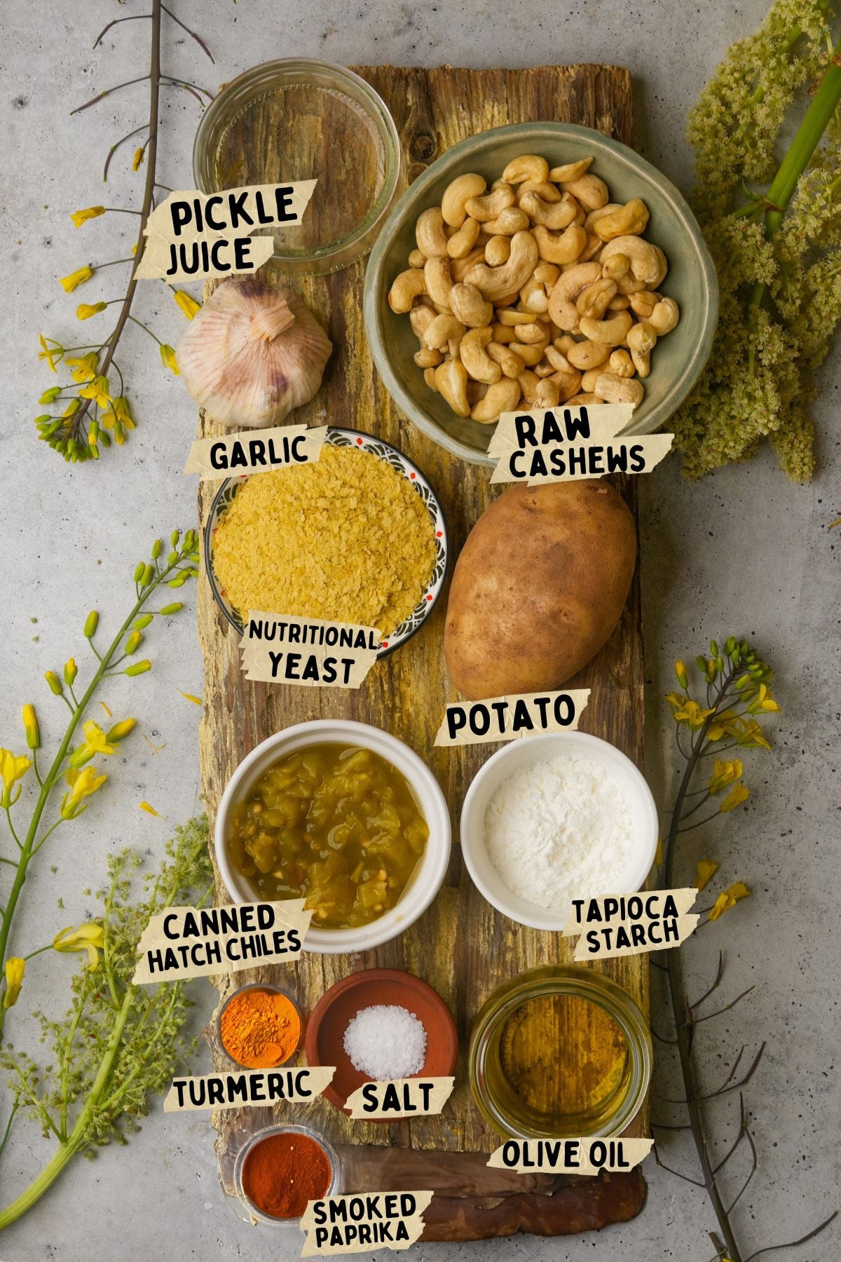 A wooden board with ingredients for a vegan queso recipe including bowls of raw cashews, nutritional yeast, canned hatch chiles, tapioca starch, olive oil, and spices, alongside a garlic bulb, potato, and other ingredients surrounded by yellow flowers.