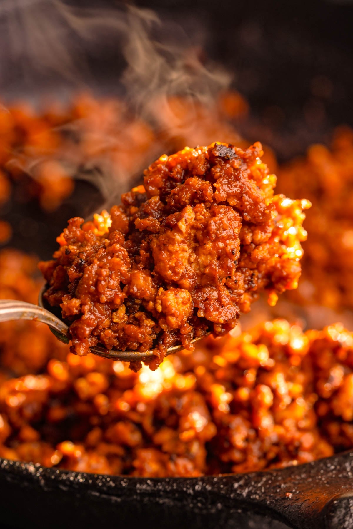 A close-up of a spoonful of steaming, seasoned vegan ground beef, held above a pan filled with more of the cooked vegan taco salad meat.