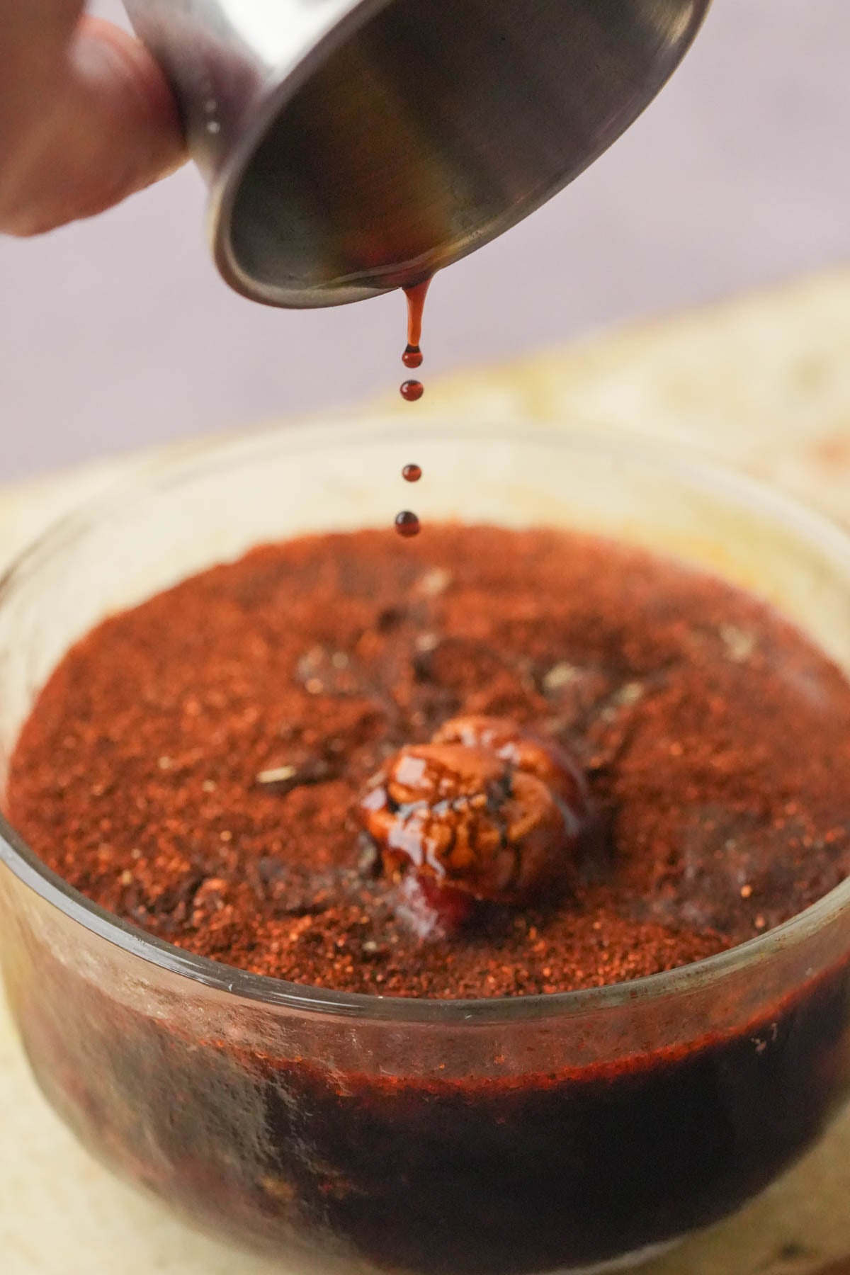 A metal cup pours tamari onto a bowl filled with a reddish-brown vegan taco meat marinade.
