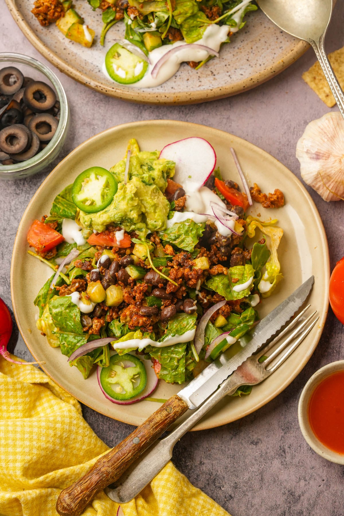 A plate of vegan taco salad with lettuce, black beans, tomatoes, radishes, guacamole, sliced jalapeños, and drizzled dressing, served with a fork and knife. A bowl of sliced black olives and another plate are nearby.