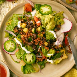 A plate of vegan taco salad with tofu ground beef, black beans, corn, lettuce, tomato, radish slices, jalapeño, red onion, guacamole, and a drizzle of sour cream. A knife rests on the plate's edge.