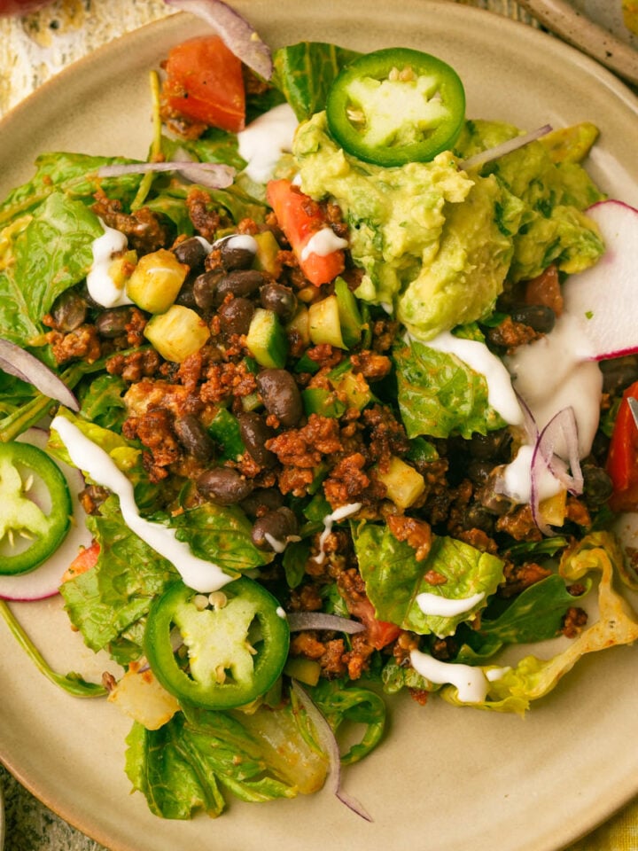 A plate of vegan taco salad with tofu ground beef, black beans, corn, lettuce, tomato, radish slices, jalapeño, red onion, guacamole, and a drizzle of sour cream. A knife rests on the plate's edge.