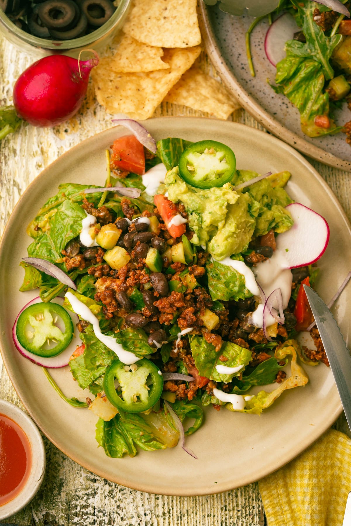 A plate of vegan taco salad with lettuce, tofu ground meat, black beans, tomatoes, corn, red onion, avocado, sliced jalapeños, radishes, a drizzle of white sauce, and guacamole. Tortilla chips and sliced olives are nearby.