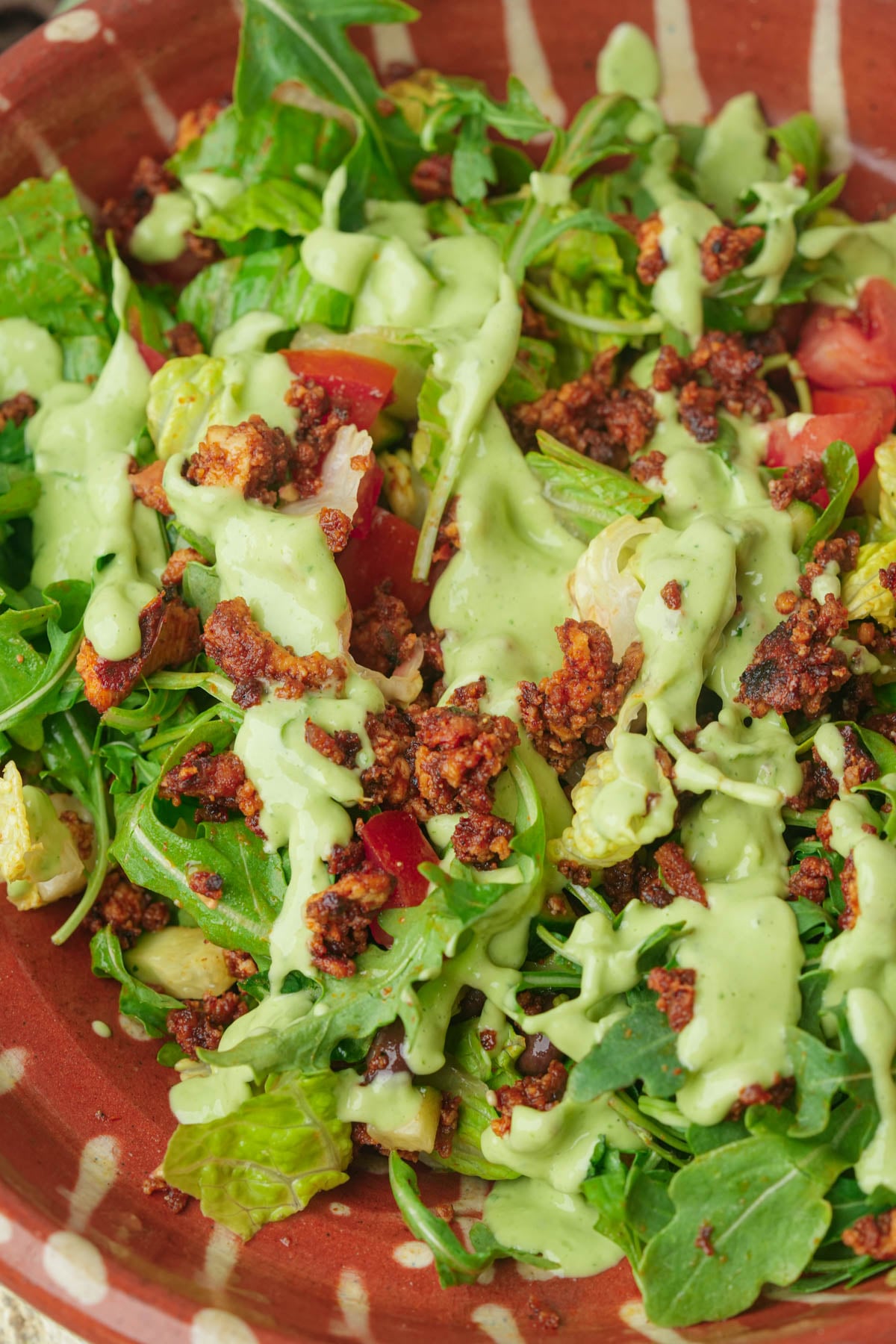 A close-up of a vegan taco salad in a brown bowl, topped with crispy crumbled tofu bits, leafy greens, diced tomatoes, and drizzled with a creamy green dressing.