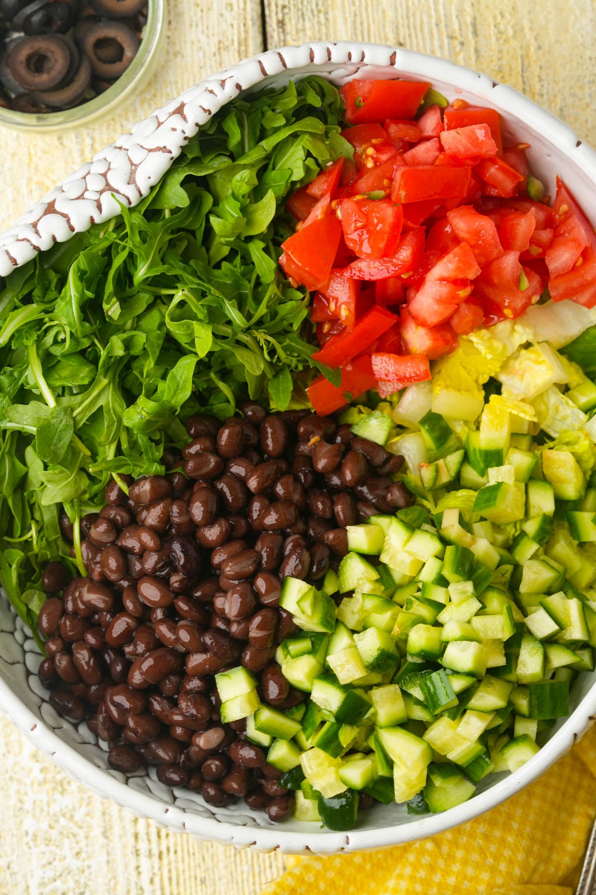 A bowl filled with chopped arugula, diced tomatoes, black beans, diced cucumbers, and diced avocado, arranged in separate sections on a light wooden surface. A small bowl of sliced black olives is in the background.