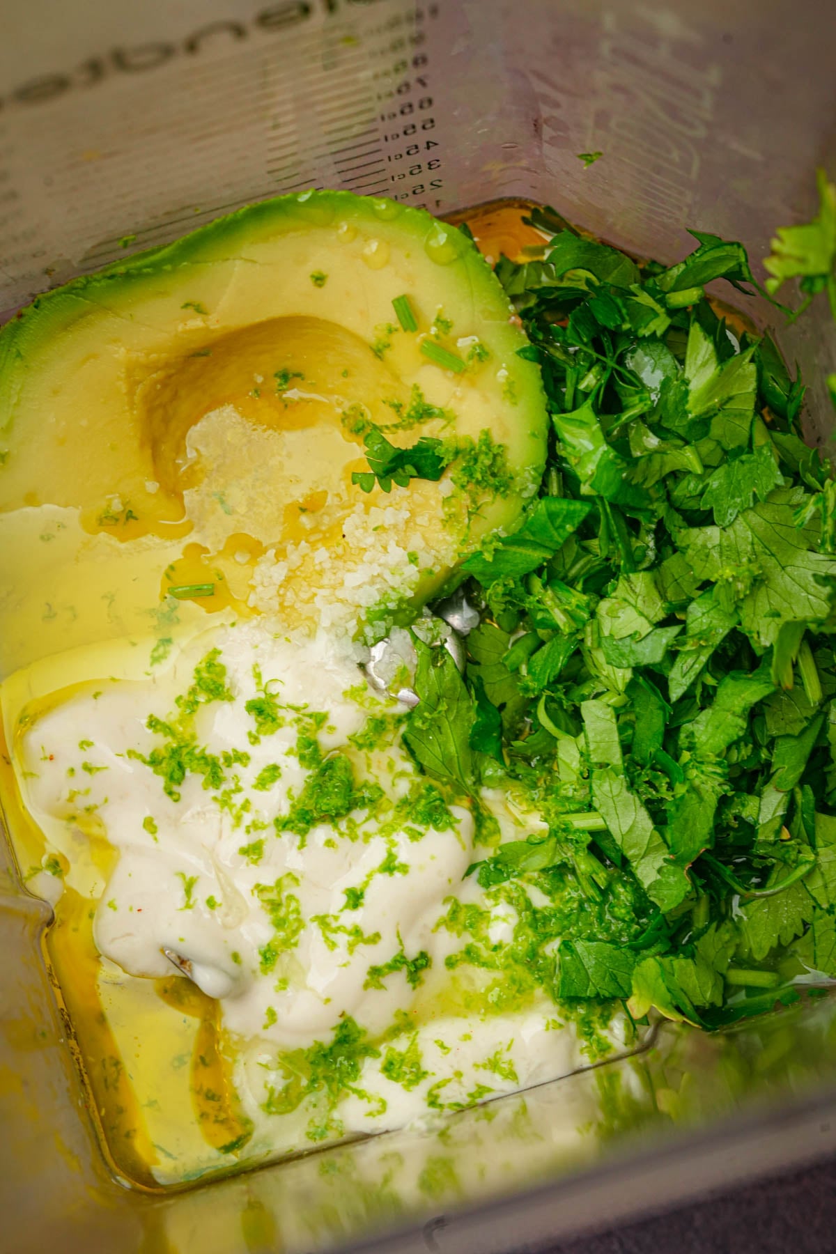 A close-up of a blender containing half an avocado, chopped cilantro, vegan sour cream, lime zest, olive oil, and coarse salt, ready to be blended into vegan taco salad dressing.