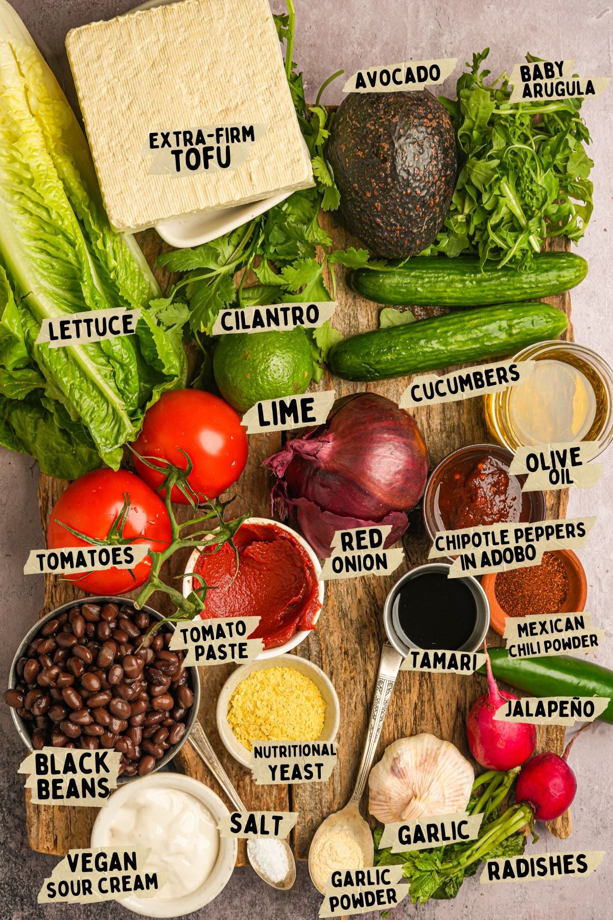 Labeled ingredients arranged on a table for a vegan taco salad recipe including tofu, avocado, arugula, lettuce, cilantro, tomatoes, lime, red onion, cucumbers, black beans, seasonings, and sauces. Labels identify each item.