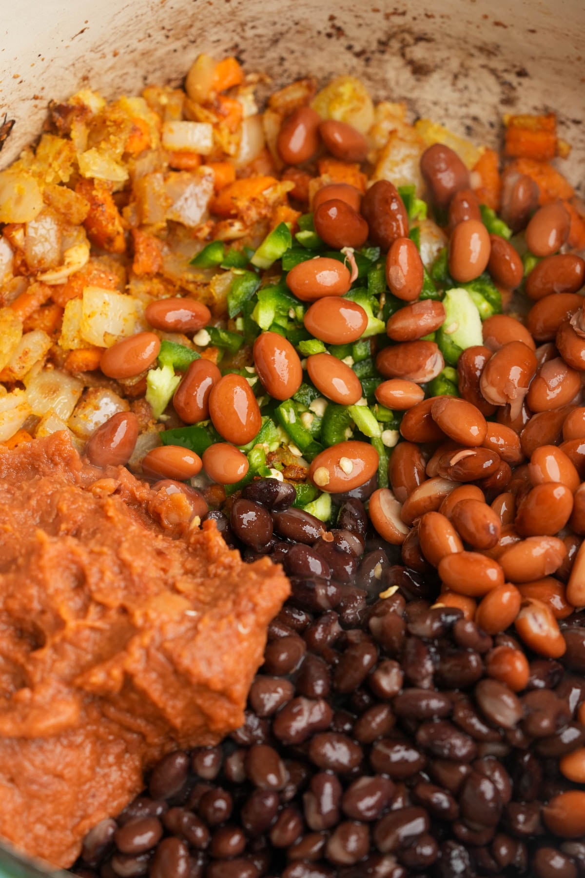 Chopped onions, diced green peppers, pinto beans, black beans, and refried beans arranged in sections in a pot, ready to be mixed together.