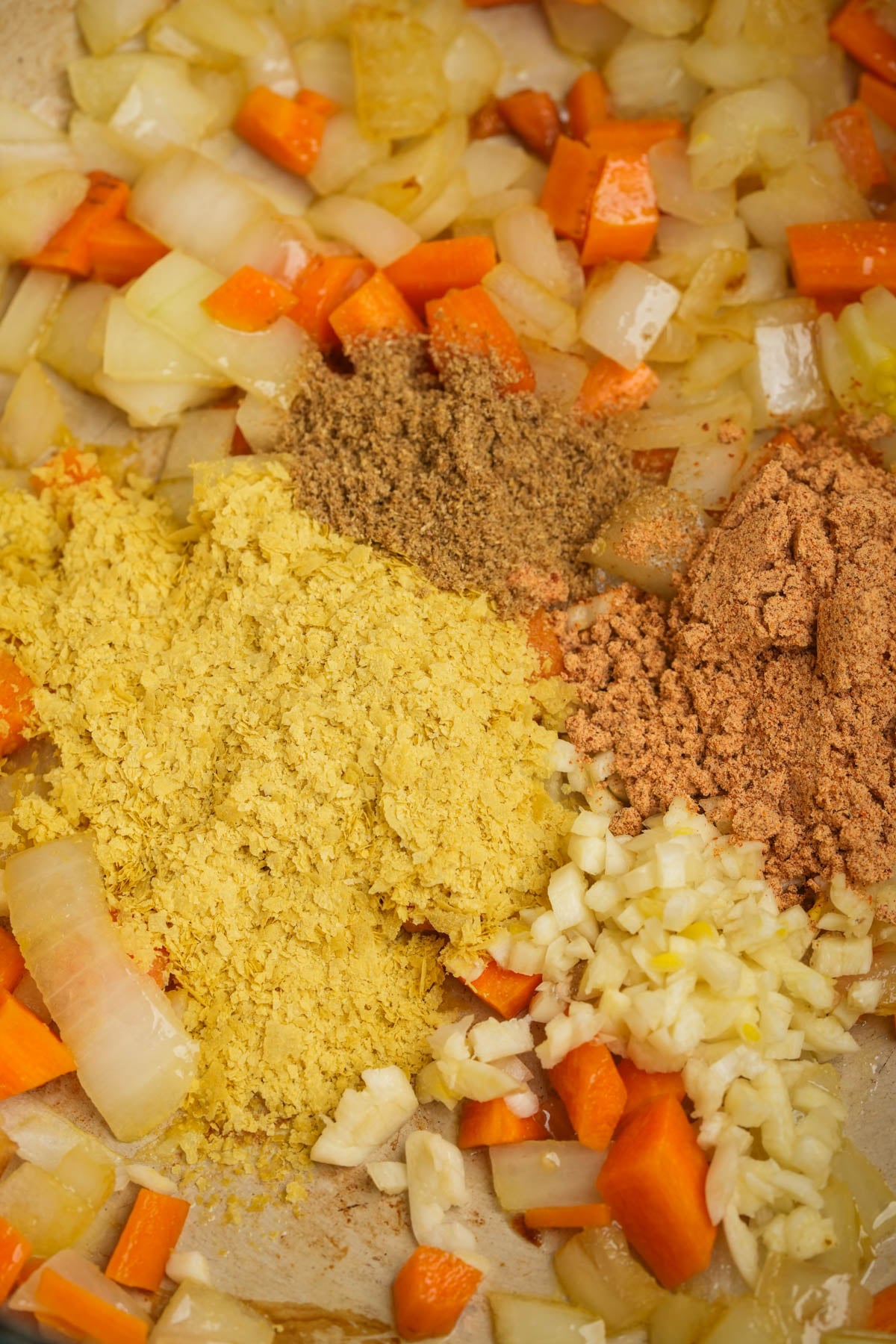 Chopped onions, carrots, and garlic cooking in a pan with piles of ground spices and nutritional yeast.