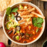 A bowl of vegan taco soup topped with avocado cubes, black olives, jalapeño slices, cilantro, sour cream, and crispy tortilla strips, placed on a rustic wooden table.
