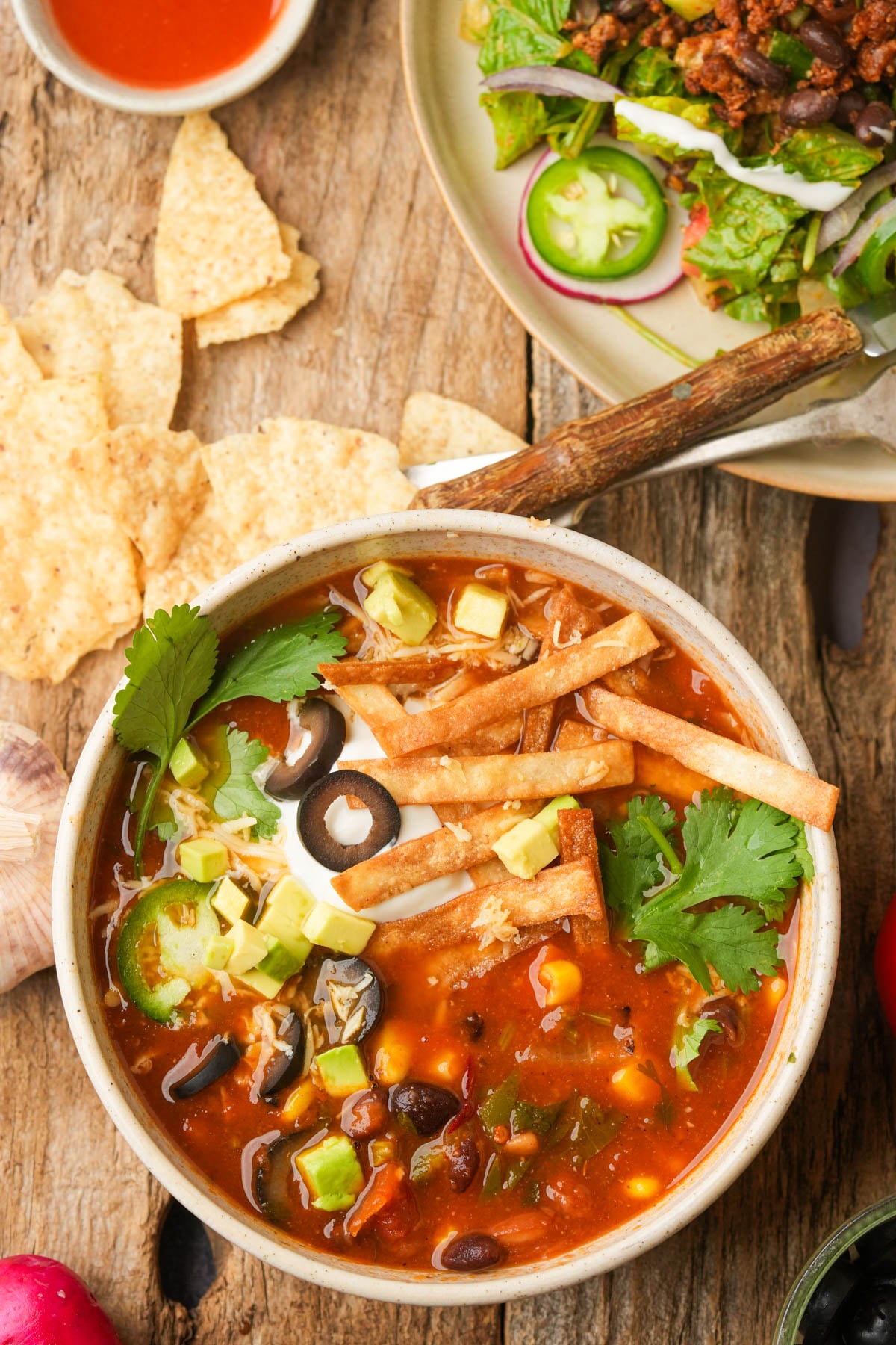 A bowl of vegan taco soup topped with sliced avocado, black olives, cilantro, tortilla strips, and a dollop of sour cream sits on a rustic wooden table beside tortilla chips and a plate of salad.
