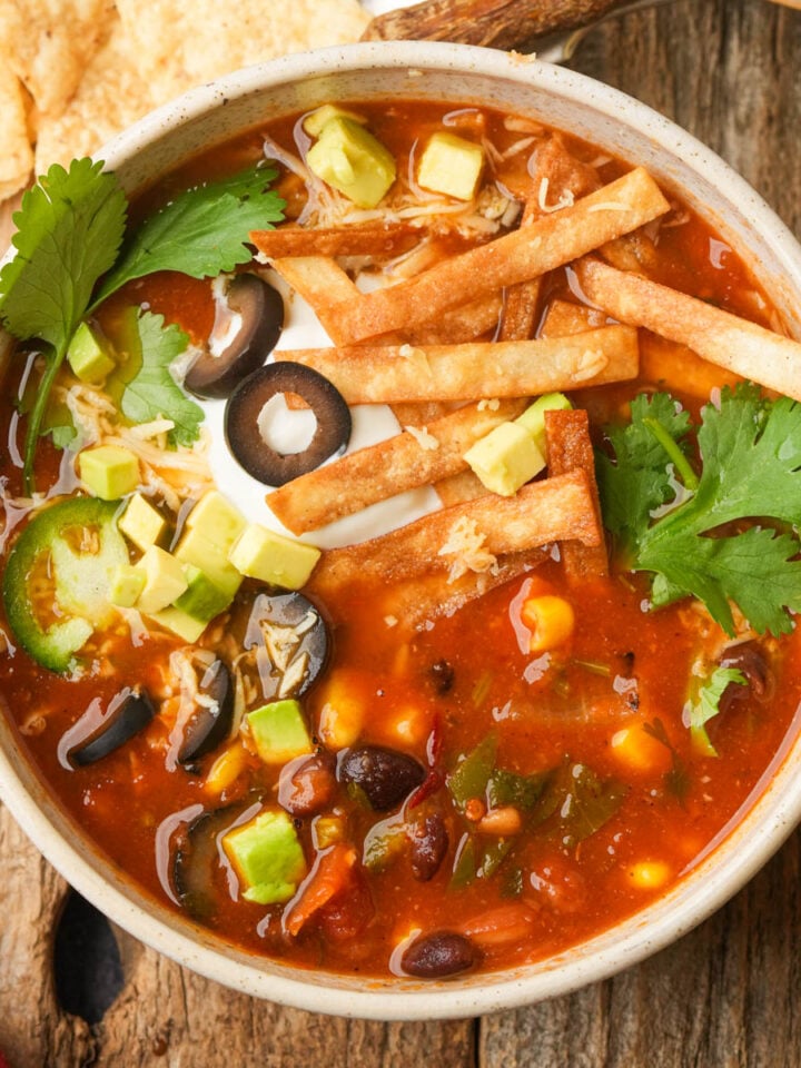 A bowl of vegan taco soup topped with avocado cubes, black olives, jalapeño slices, cilantro, sour cream, and crispy tortilla strips, placed on a rustic wooden table.