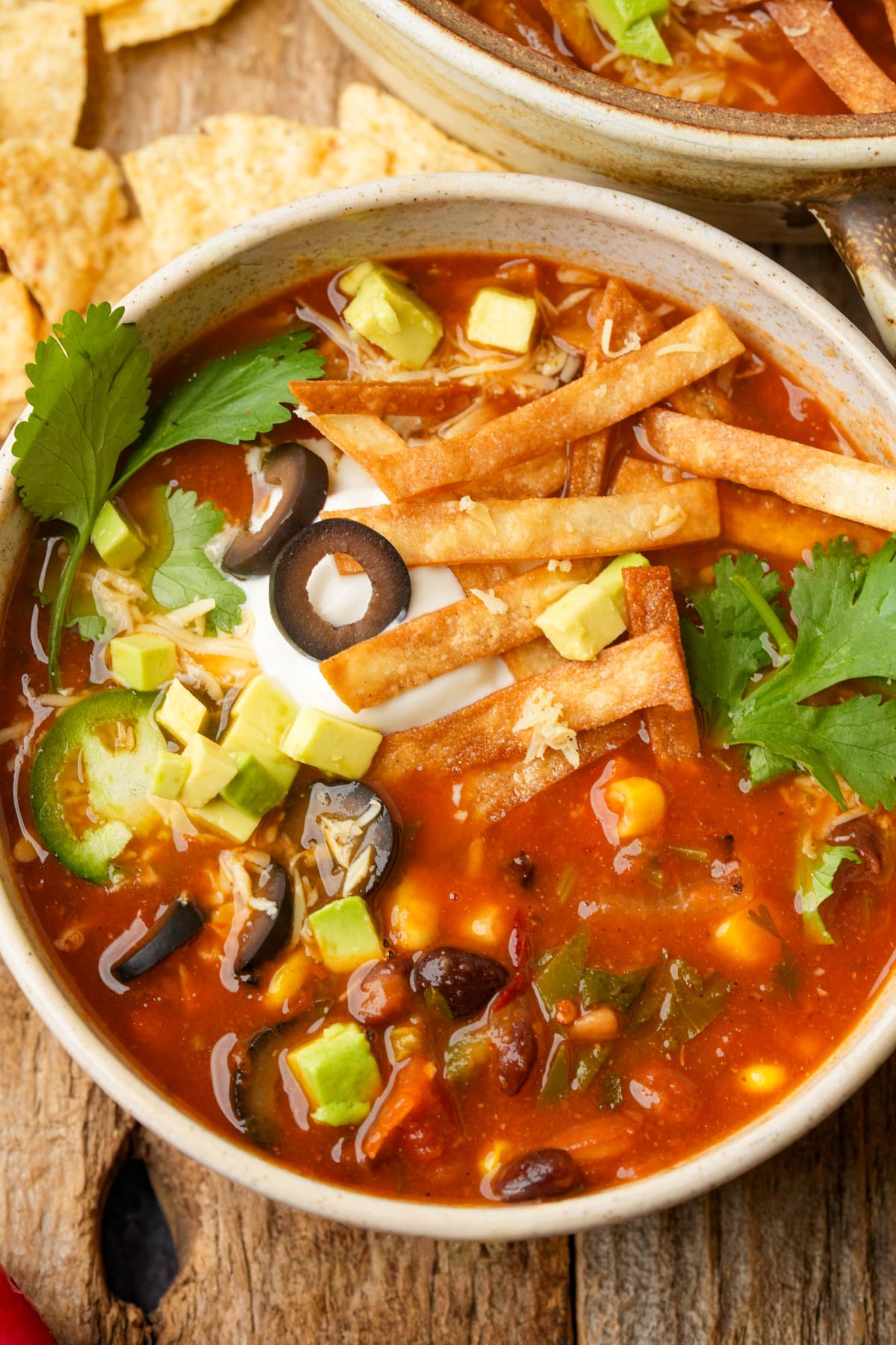 A bowl of vegan taco soup topped with avocado cubes, sliced black olives, jalapeños, cilantro, crispy tortilla strips, and a dollop of sour cream, with tortilla chips nearby on a rustic wooden surface.
