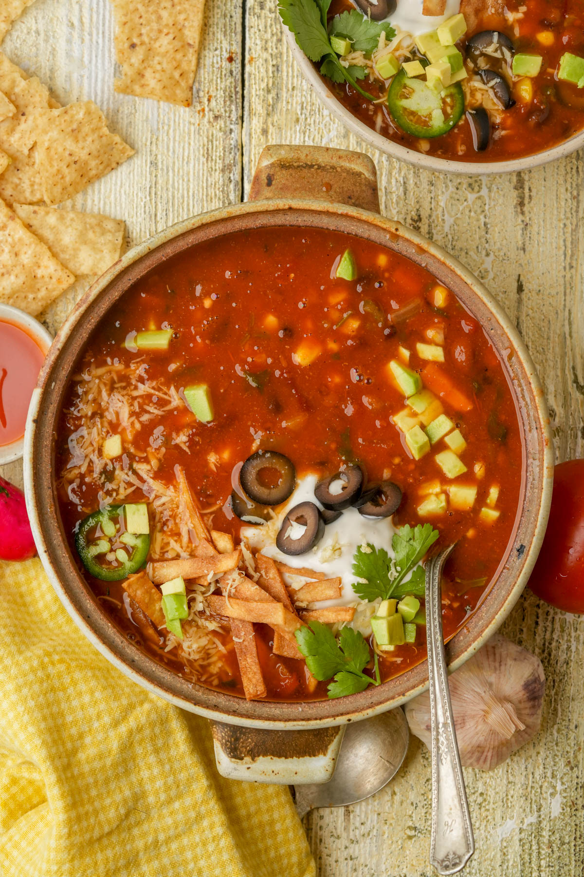 A bowl of vegan taco soup topped with shredded cheese, avocado cubes, sliced black olives, jalapeños, sour cream, cilantro, and tortilla strips, with a side of tortilla chips on a rustic wooden table.
