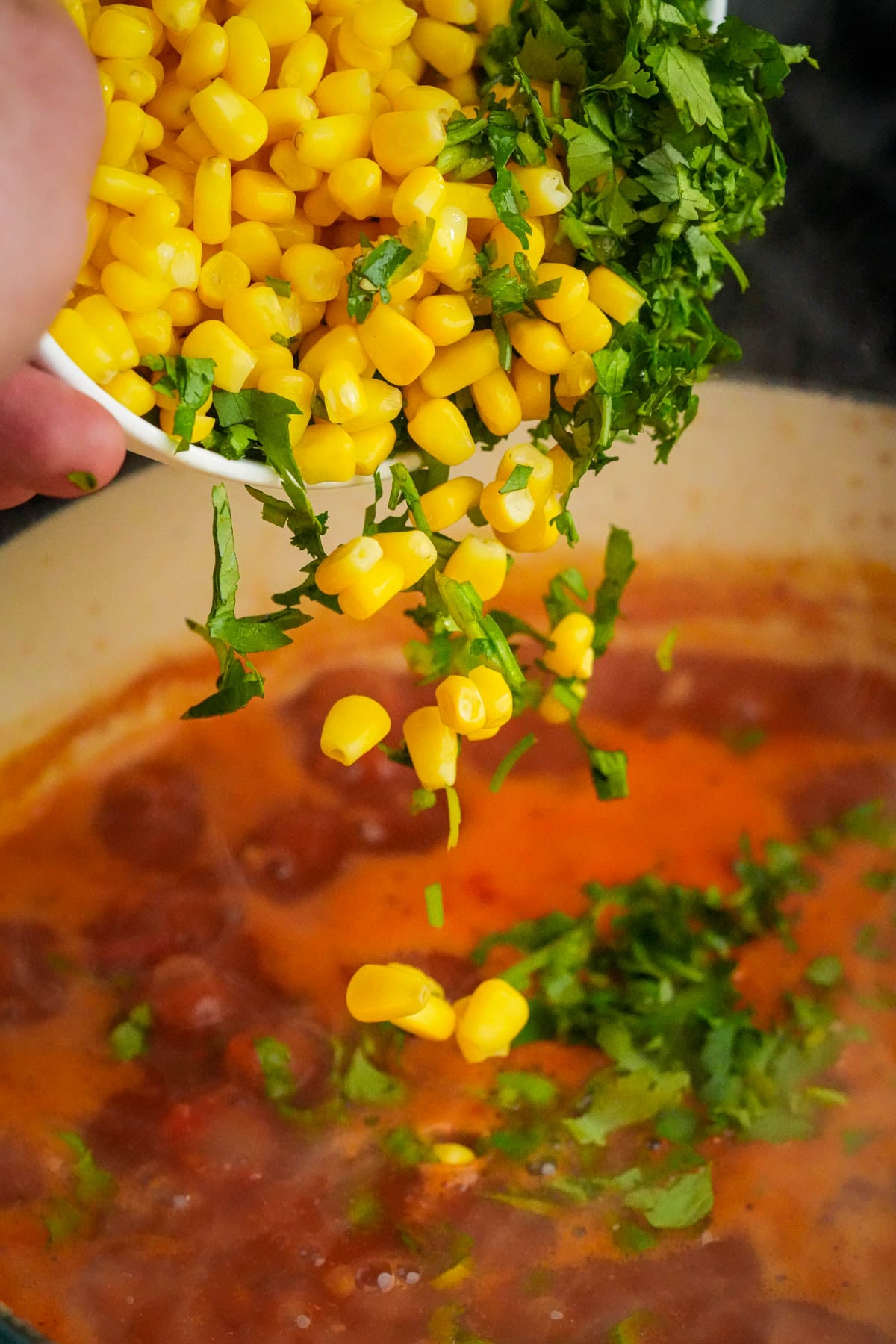A hand pours yellow corn and chopped cilantro from a bowl into a pot of simmering vegan taco soup.